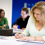 Adult student studies at desk with two classmates talking behind her.